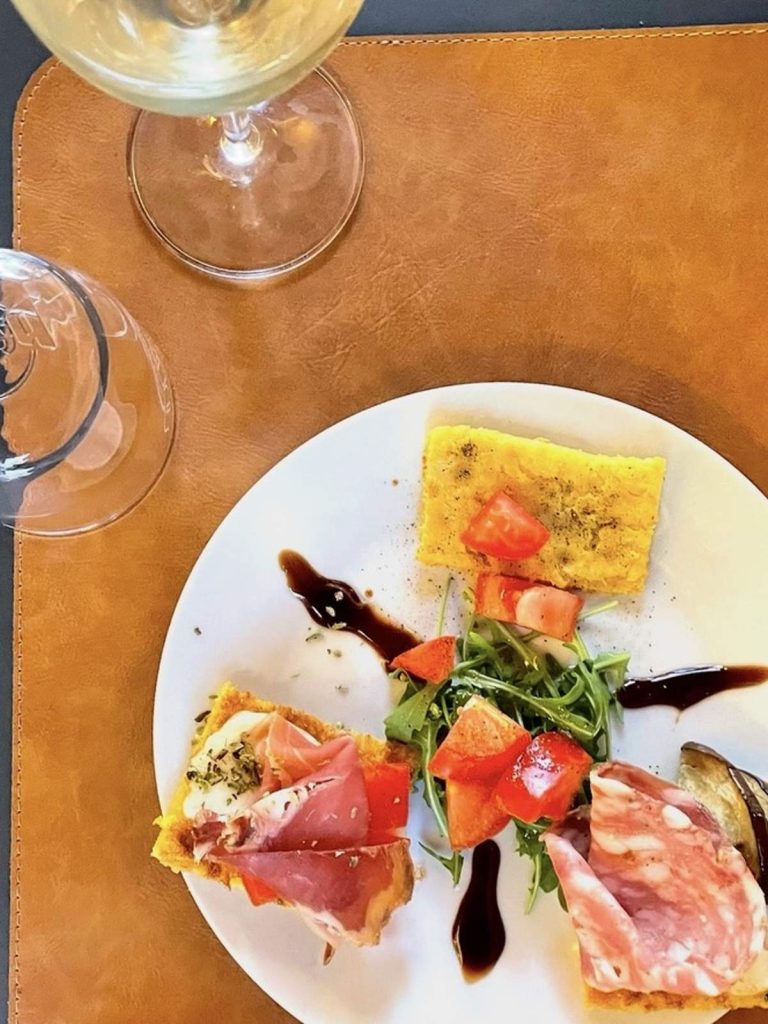 A tasting plate with three cecina squares topped with prosciutto, salami and herbs, served with arugula, tomato and a glass of white wine